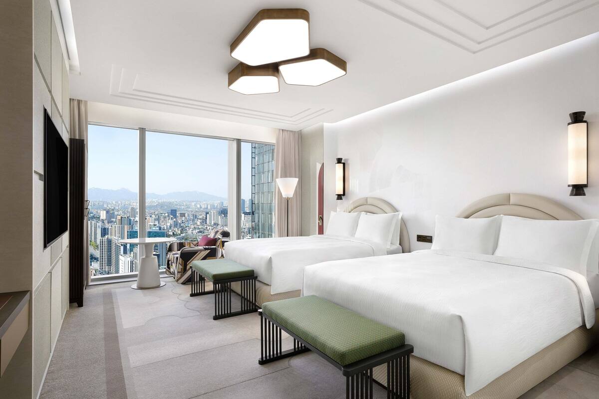 Habitación de hotel con dos camas, un ventanal con vistas a la ciudad, un televisor de pantalla plana y una pequeña mesa con un banco.