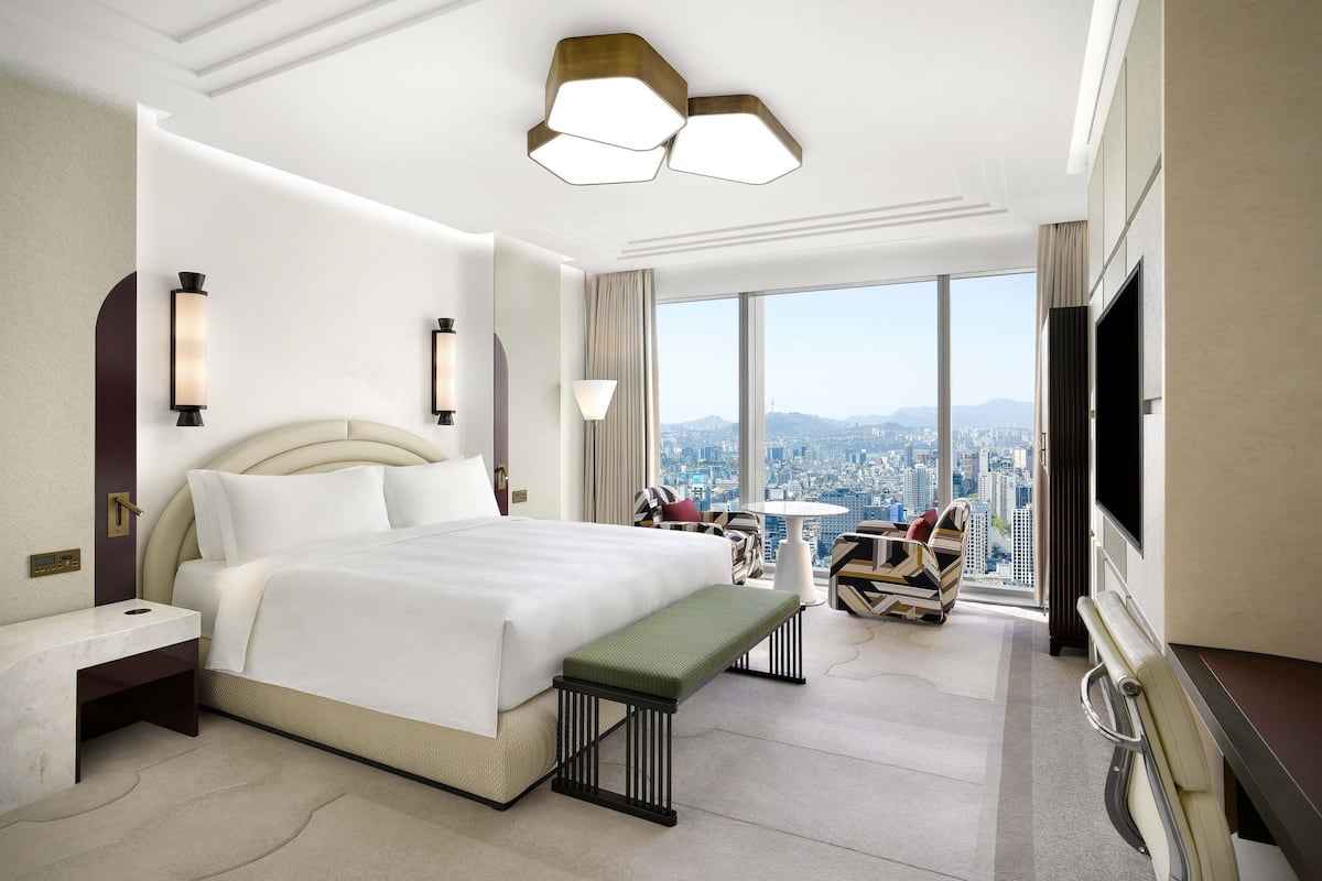 Habitación de hotel moderna con una cama grande, un banco, un escritorio, un televisor y vistas a la ciudad.