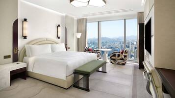 A modern hotel room with a large bed, a bench, a desk, a TV, and a city view.