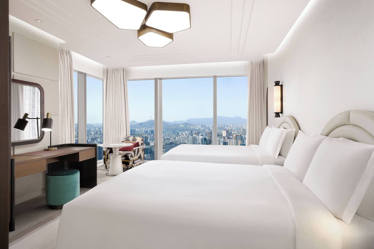 Habitación de hotel con dos camas, un escritorio, una silla y un ventanal con vistas a la ciudad.