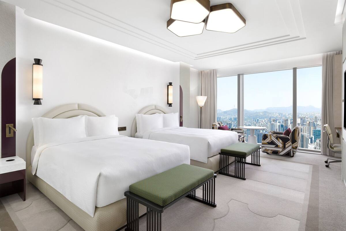 Habitación de hotel con dos camas, un banco, un escritorio y un ventanal con vistas a la ciudad.