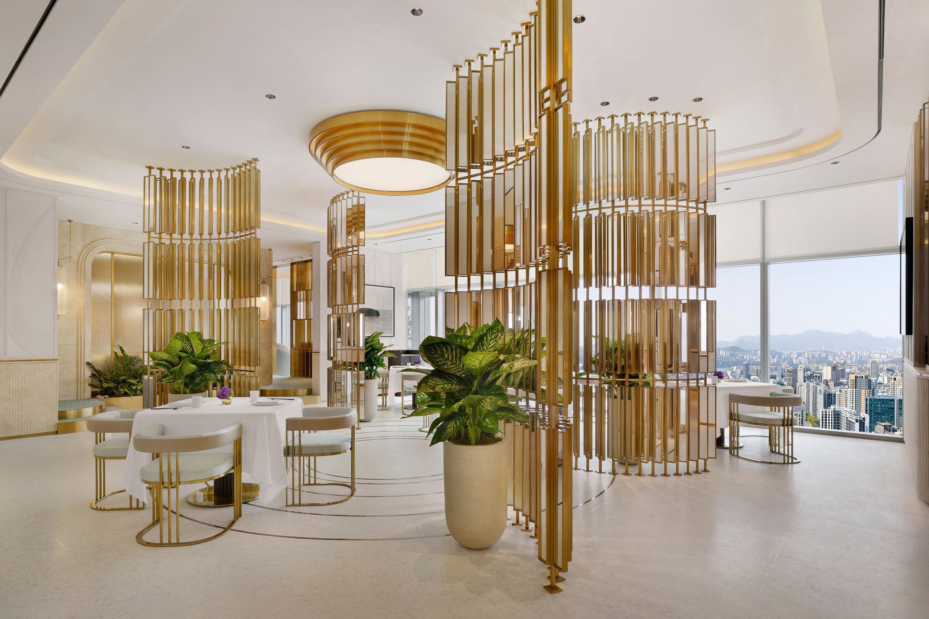 A modern hotel lobby with a high ceiling, large windows offering a city view, and a central seating area with tables and chairs.