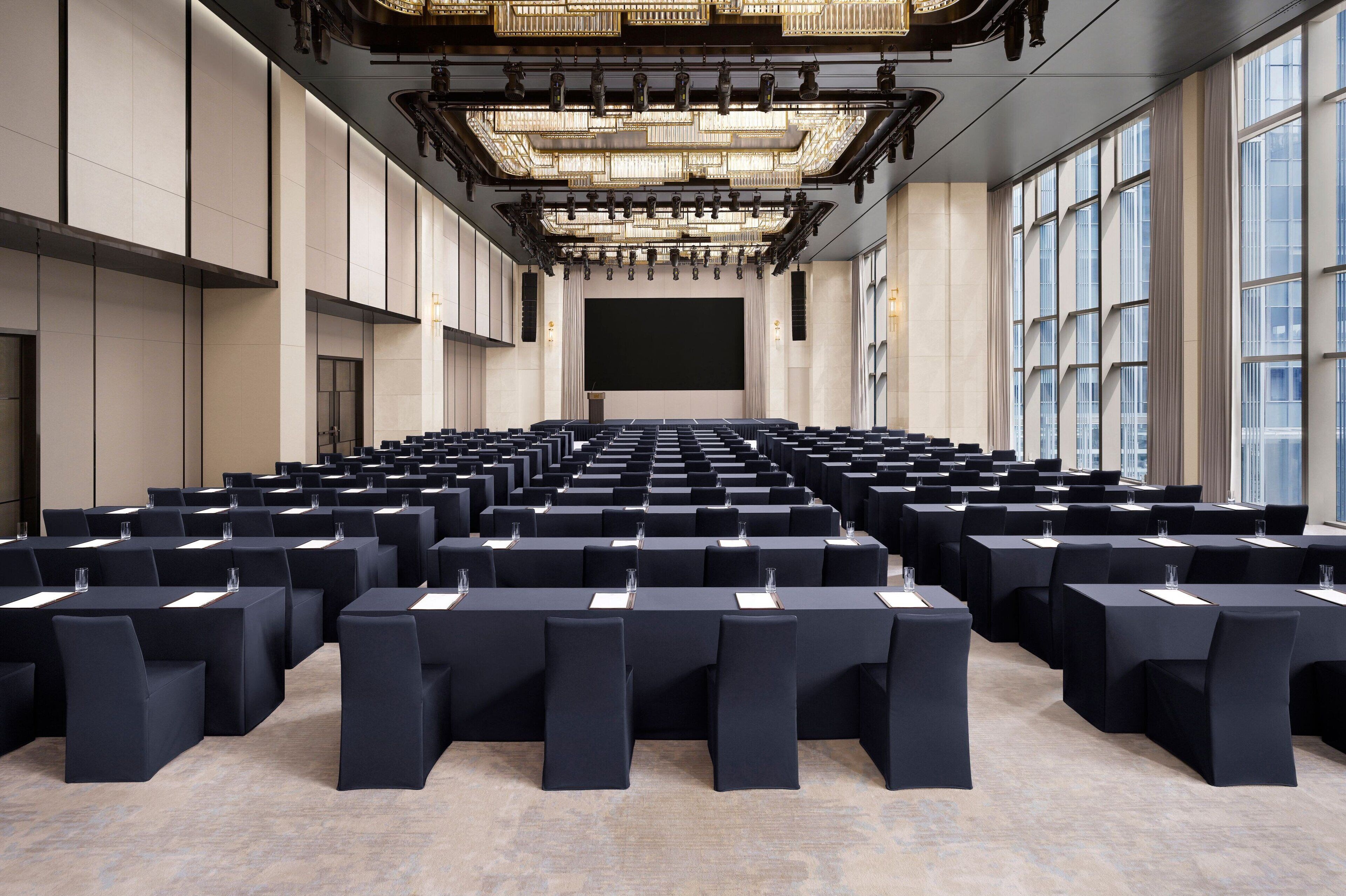 A large conference room with rows of tables and chairs, a large screen at the front, and a chandelier overhead.