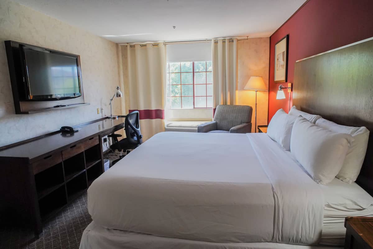 A hotel room with a large bed, a TV mounted on the wall, a desk with a chair, and a window with curtains.