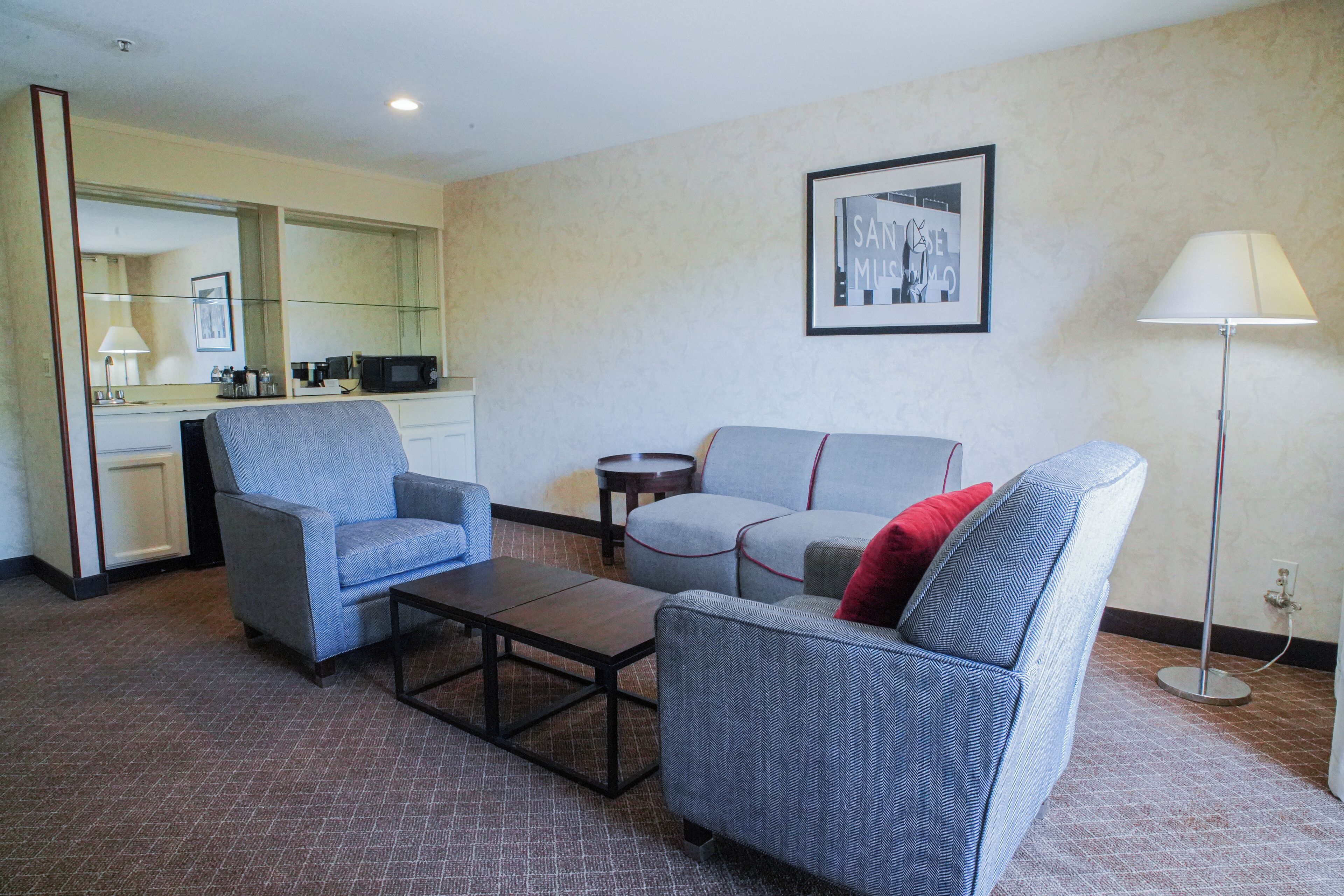 A hotel room with a sofa, armchair, coffee table, and a small side table. There is a lamp, a framed picture on the wall, and a mirror above the sofa.