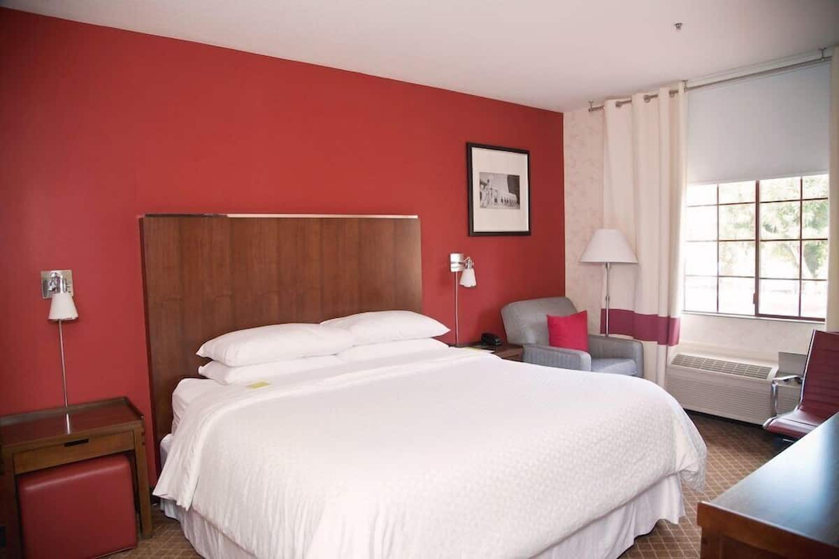 A hotel room with a large bed, a desk, a chair, a lamp, a picture on the wall, and a window with curtains.