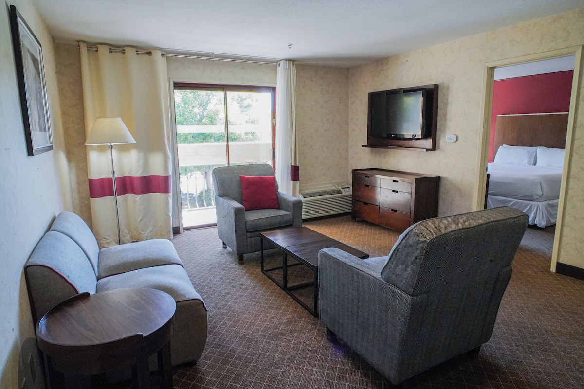 A hotel room with a sofa, armchair, a small table, a television, and a bed.