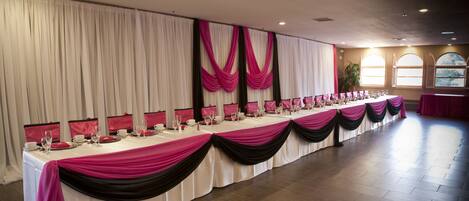 A long banquet table with white tablecloths and pink and black drapery, set up in a spacious room with wooden flooring and large windows.