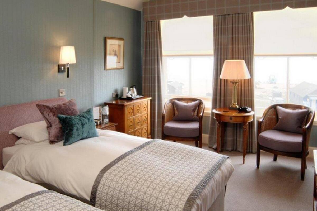 A hotel room with a large bed, two armchairs, a small table, and a lamp. There is a window with curtains and a view of the city.