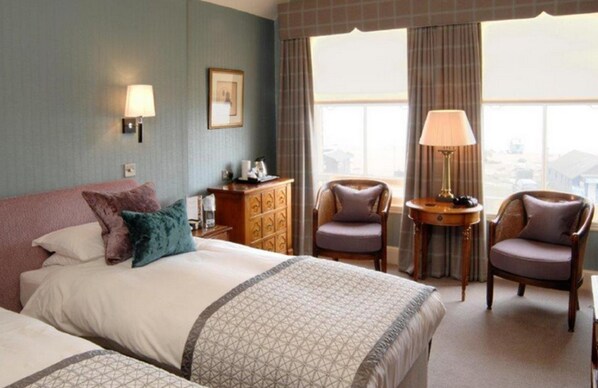 A hotel room with a large bed, two armchairs, a small table, and a lamp. There is a window with curtains and a view of the city.