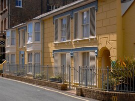 A row of yellow buildings with white shutters, a metal fence, and a brick base.