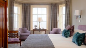 A hotel room with a large bed, two armchairs, a small table with a lamp, and a view of the city through large windows. - Wentworth Hotel (Aldeburgh)