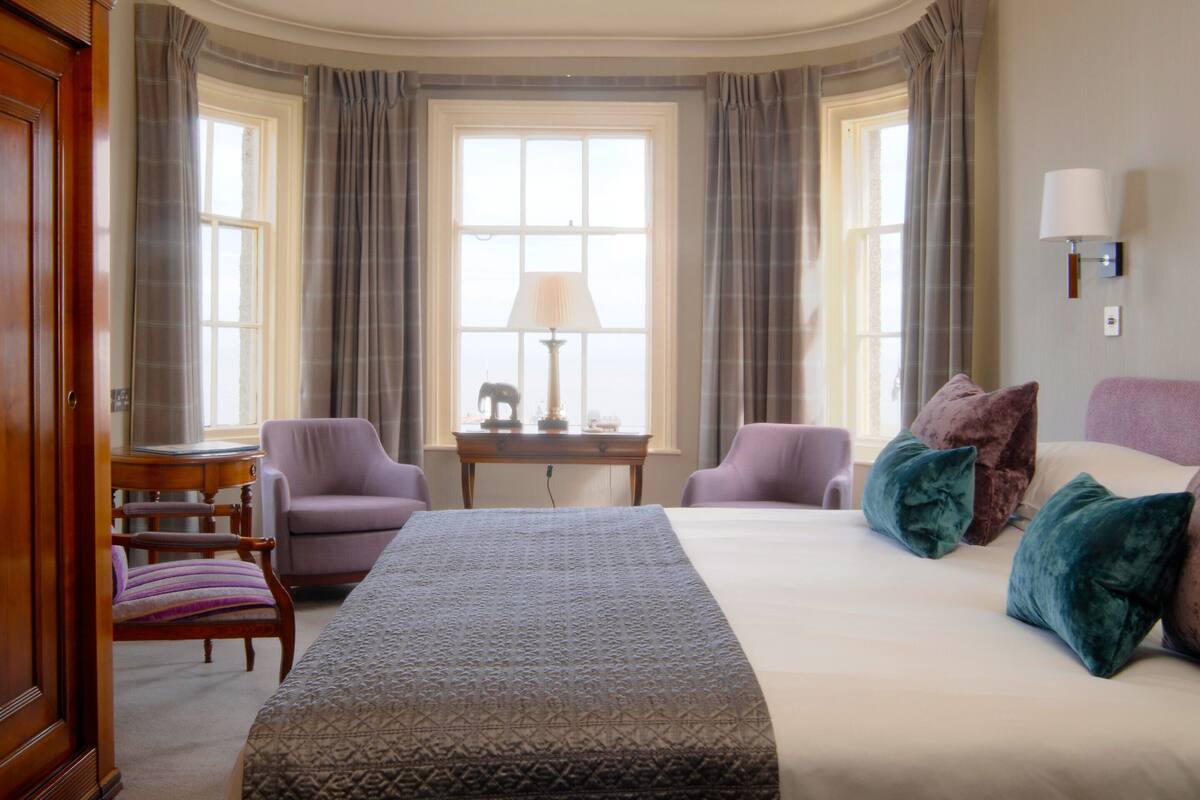 A hotel room with a large bed, two armchairs, a small table with a lamp, and a view of the city through large windows.