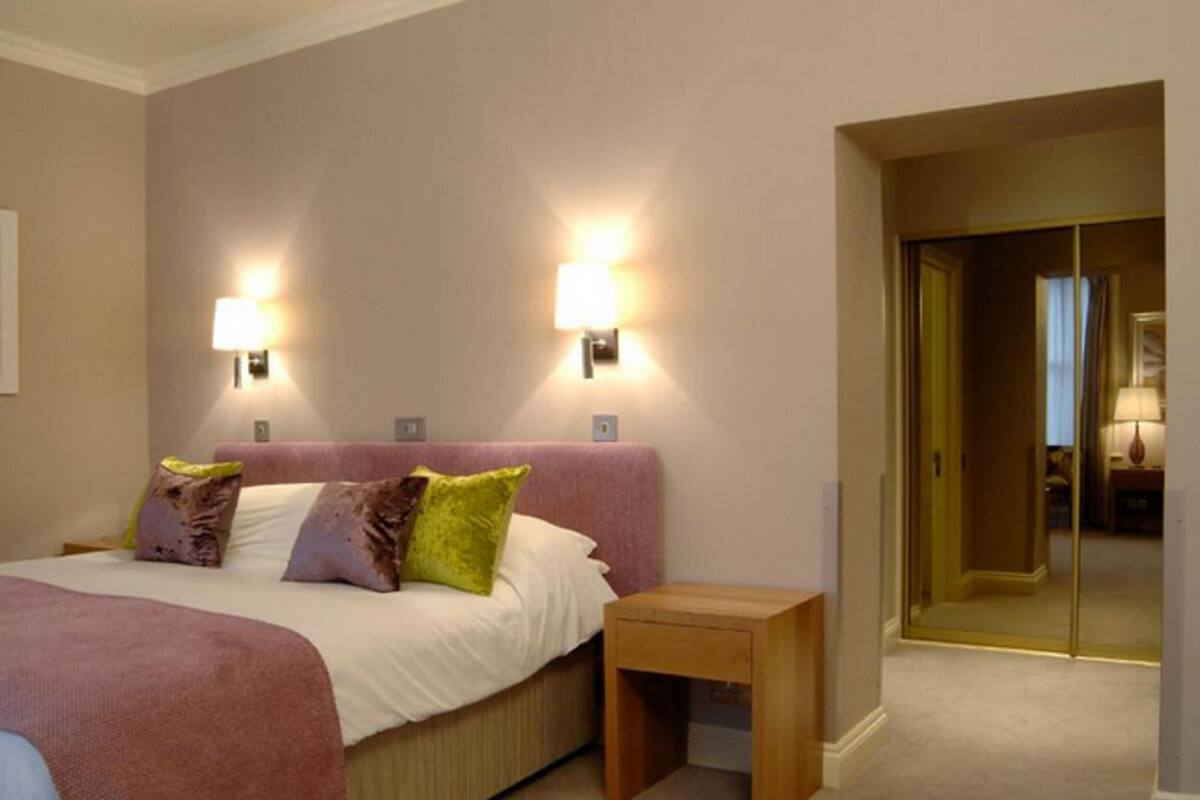 A hotel room with a large bed, two bedside lamps, a small wooden nightstand, and a door leading to another room.