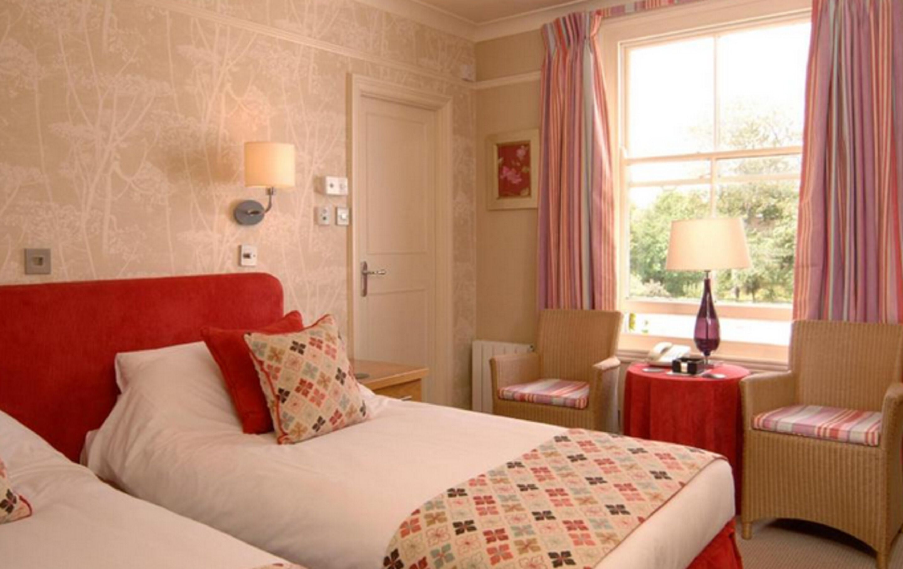 A hotel room with a large bed, two chairs, a small table with a lamp, and a window with curtains.