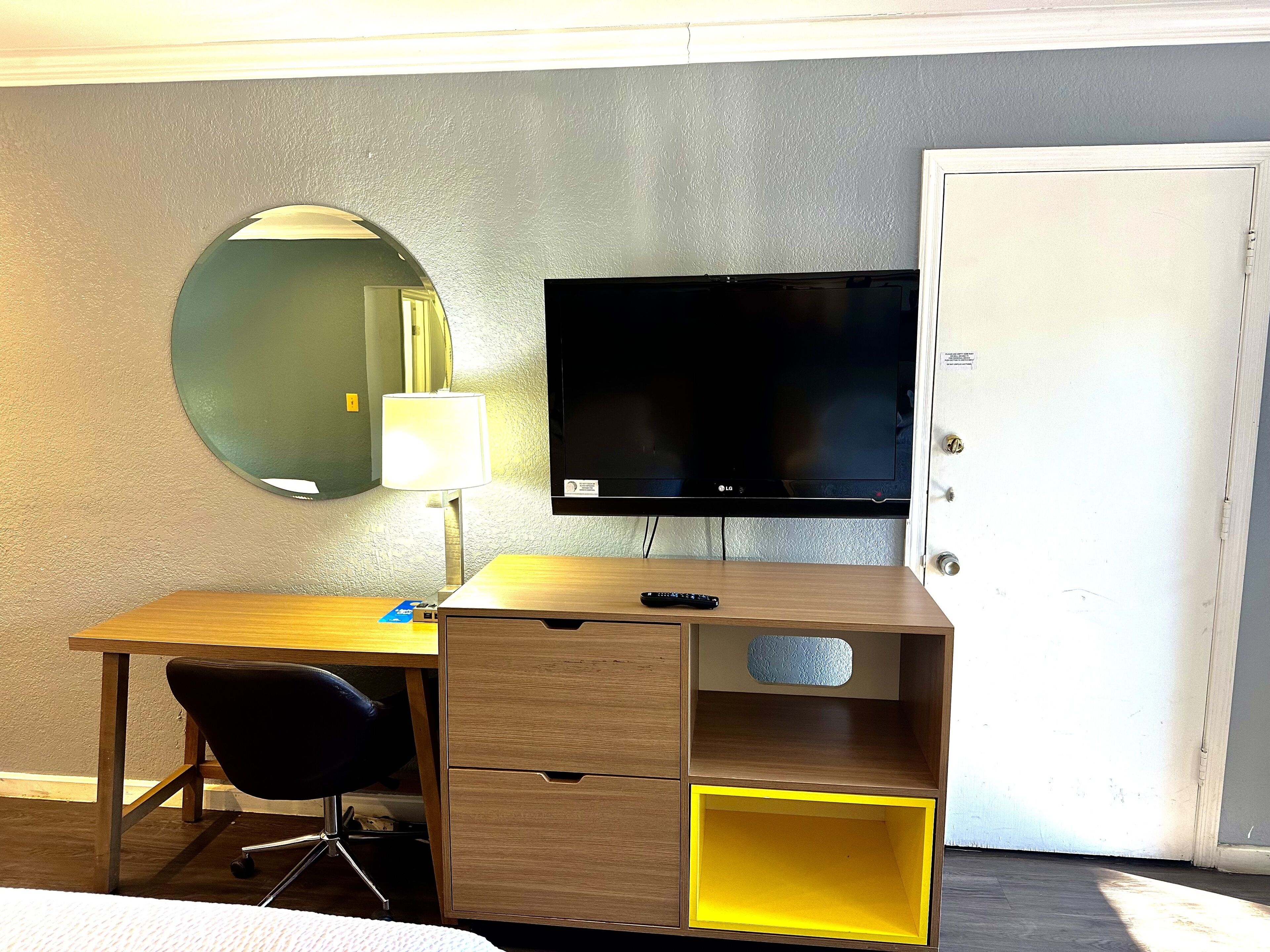 A hotel room with a wooden desk, a black chair, a flat-screen TV, a mirror, a lamp, and a door.