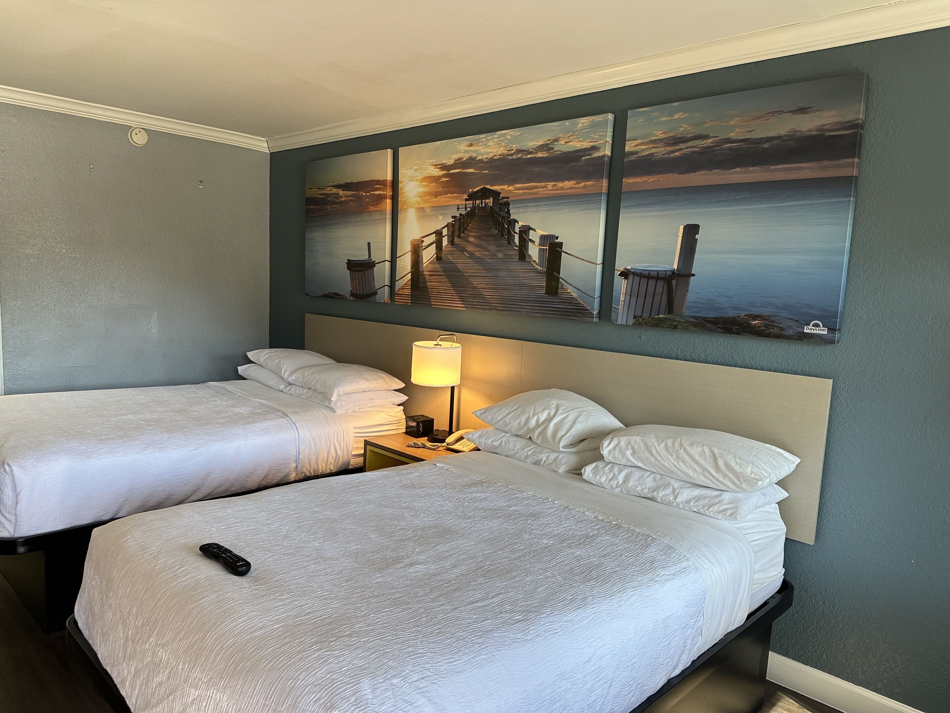 A hotel room with two single beds, a nightstand with a lamp, and a painting of a pier and sunset on the wall.