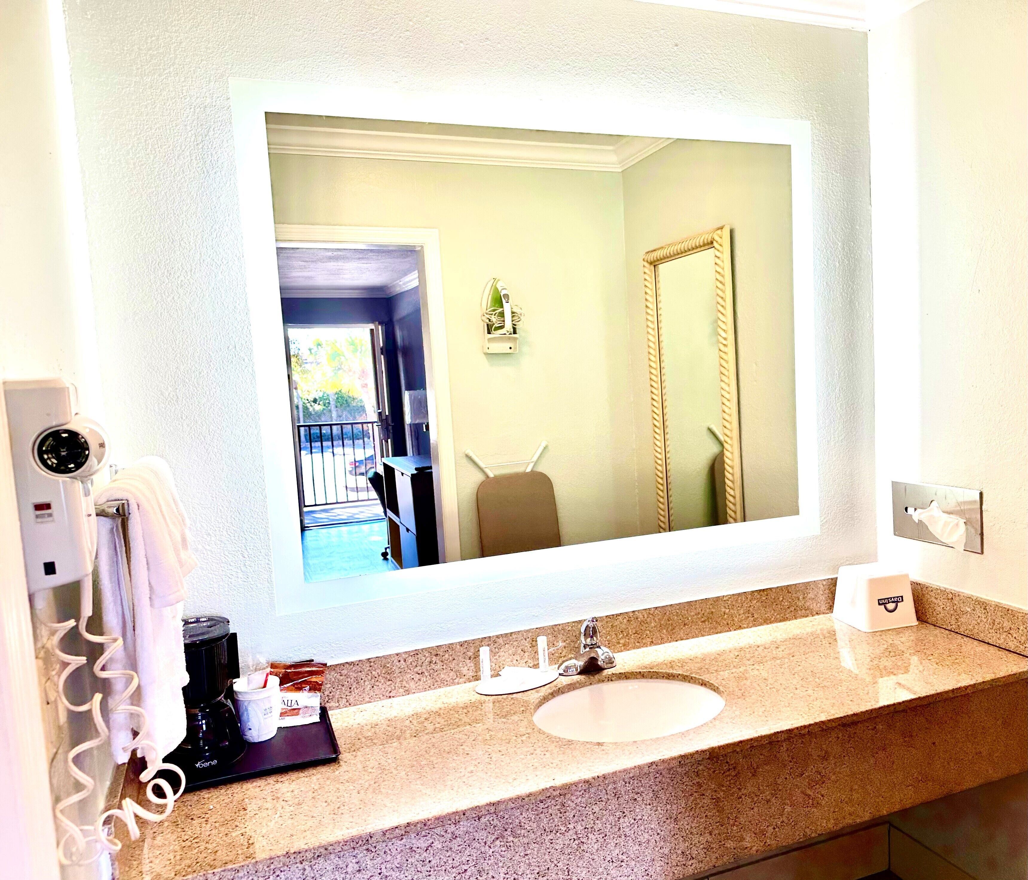A bathroom with a large mirror, a sink, a towel rack, a hairdryer, and a coffee maker.