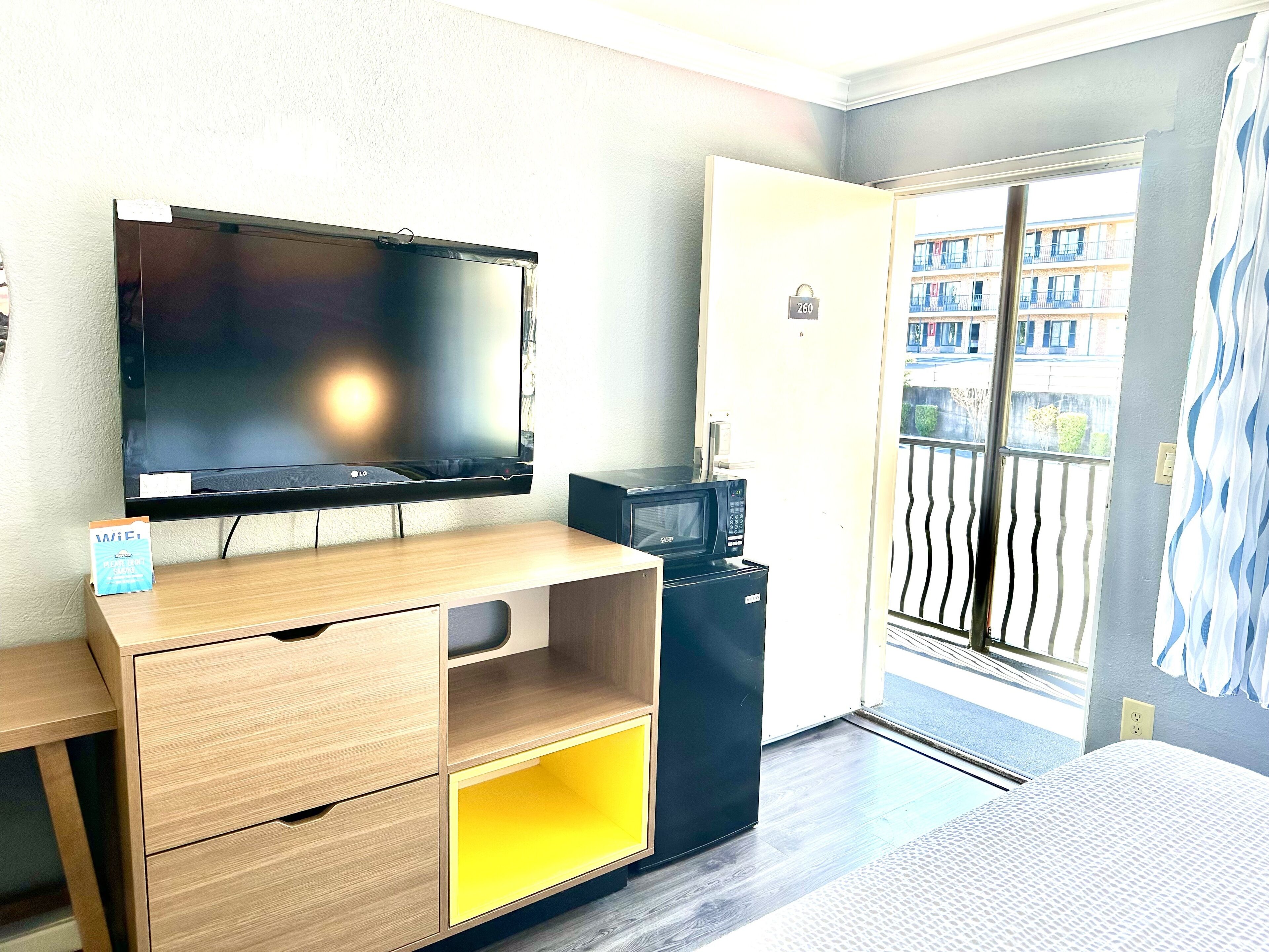 Et værelse med et fladskærms-TV, en mikrobølgeovn, et lille køleskab, et træskrivebord med skuffer og en dør, der fører ud til en balkon med udsigt over bygninger.