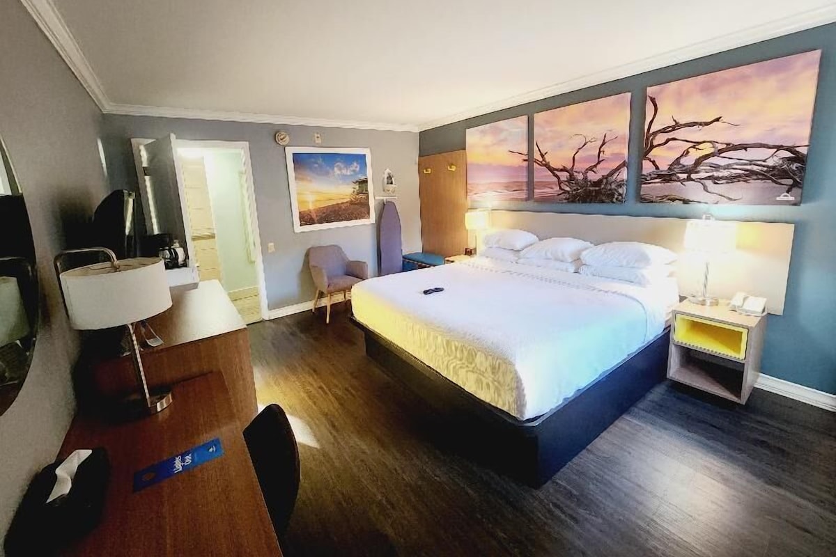 A hotel room with a bed, bedside tables, a desk, a chair, and artwork on the walls.