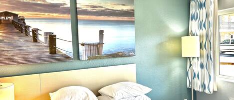 A hotel room with a bed, a nightstand with a phone, a lamp, a wall-mounted air conditioner, and a view of the sea through a window.