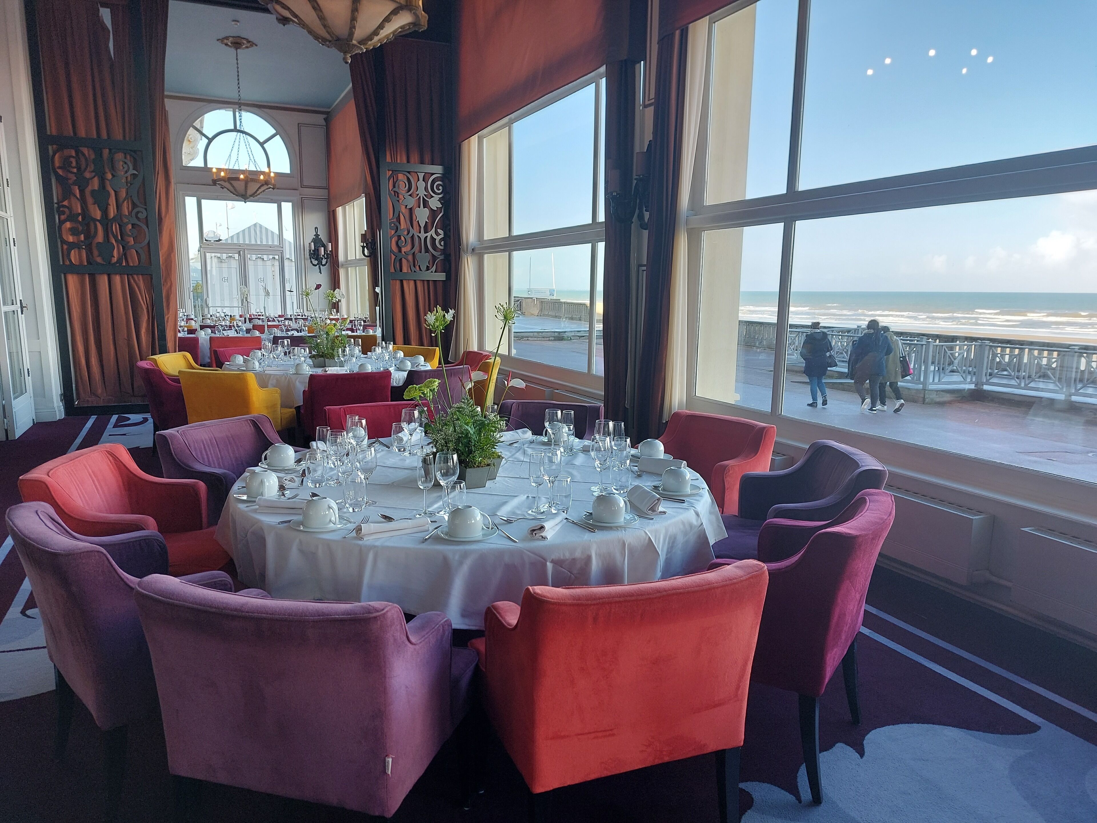 A dining area with round tables set for a meal, large windows with a view of the beach, and a balcony with people walking.