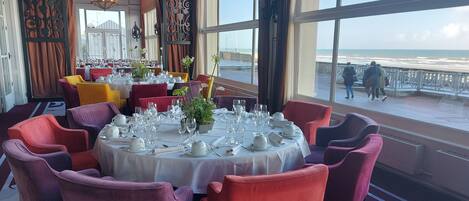 Ein Essbereich mit runden Tischen, gedeckt für eine Mahlzeit, große Fenster mit Blick auf den Strand und ein Balkon mit Passanten.