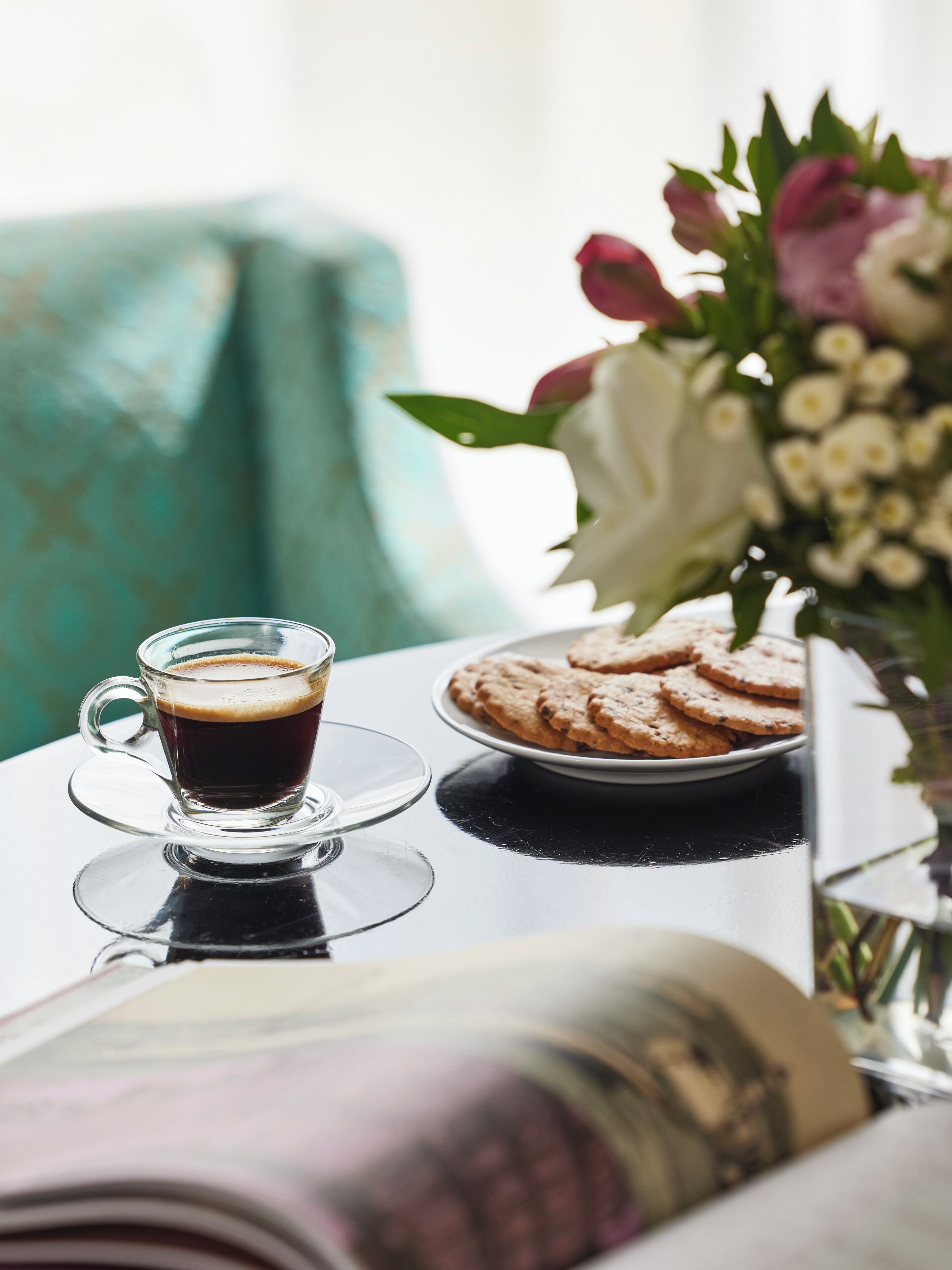 Eine transparente Tasse Kaffee auf einem Untertasse, ein Teller mit Keksen und eine Vase mit Blumen auf einem Tisch.
