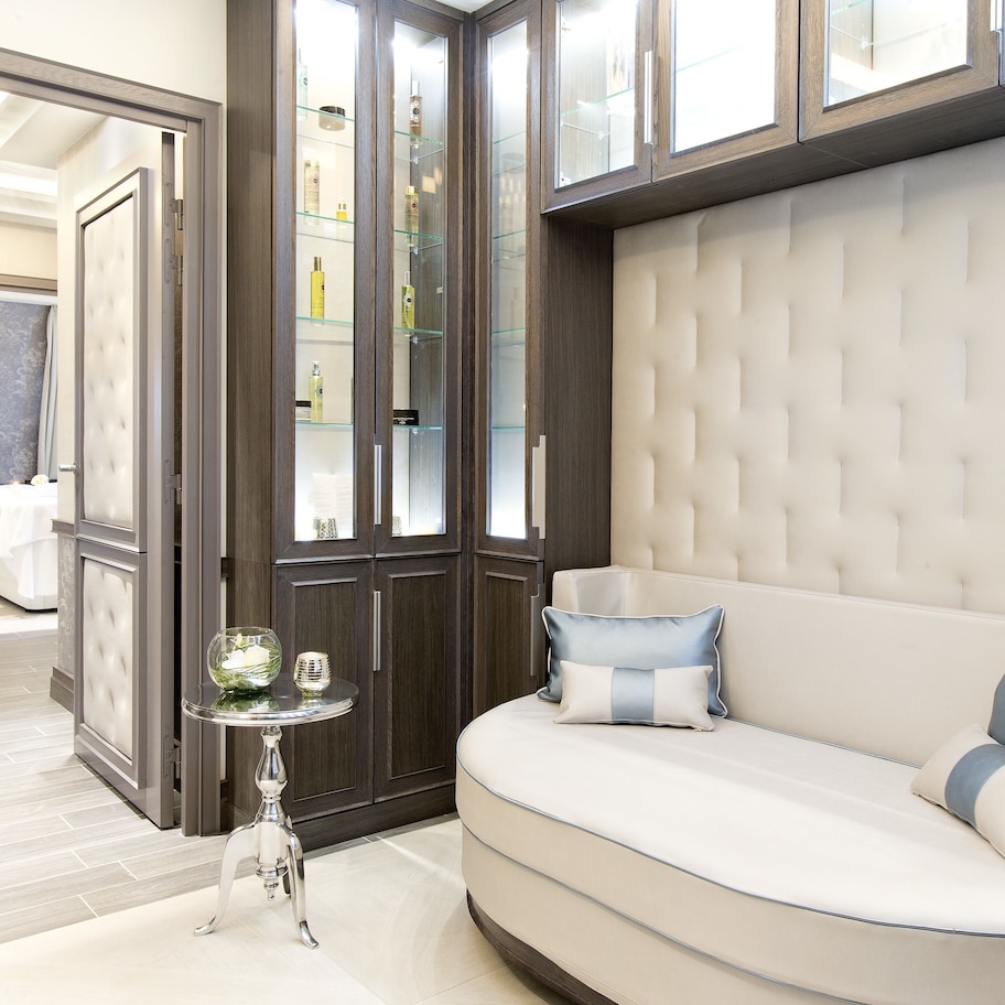 A modern bedroom with a curved sofa, a glass cabinet with toiletries, and a chandelier.