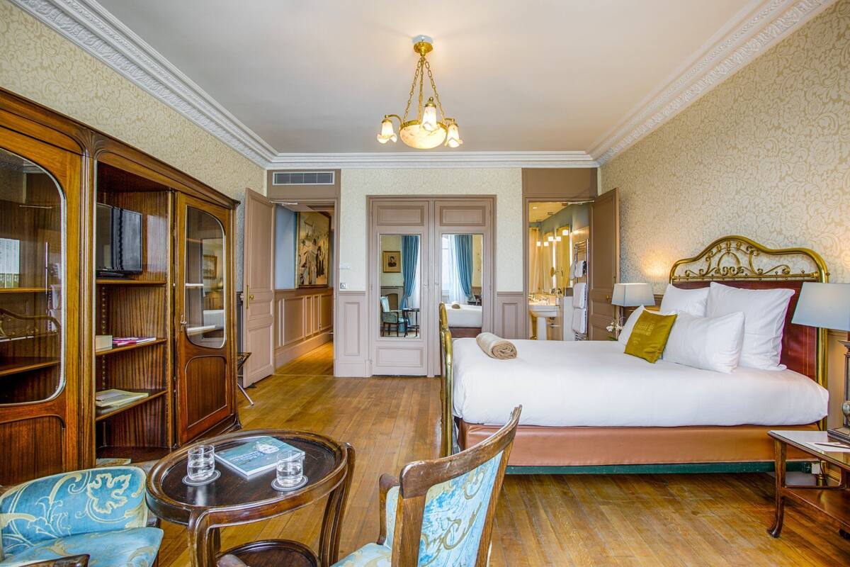 A hotel room with a large bed, a wooden wardrobe, a sitting area with a table and chairs, and a bathroom visible through an open door.