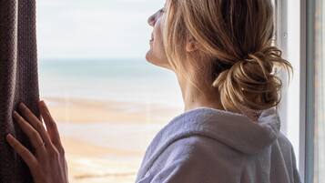 Una persona vestida con bata apoyada en una ventana con vista a la playa.