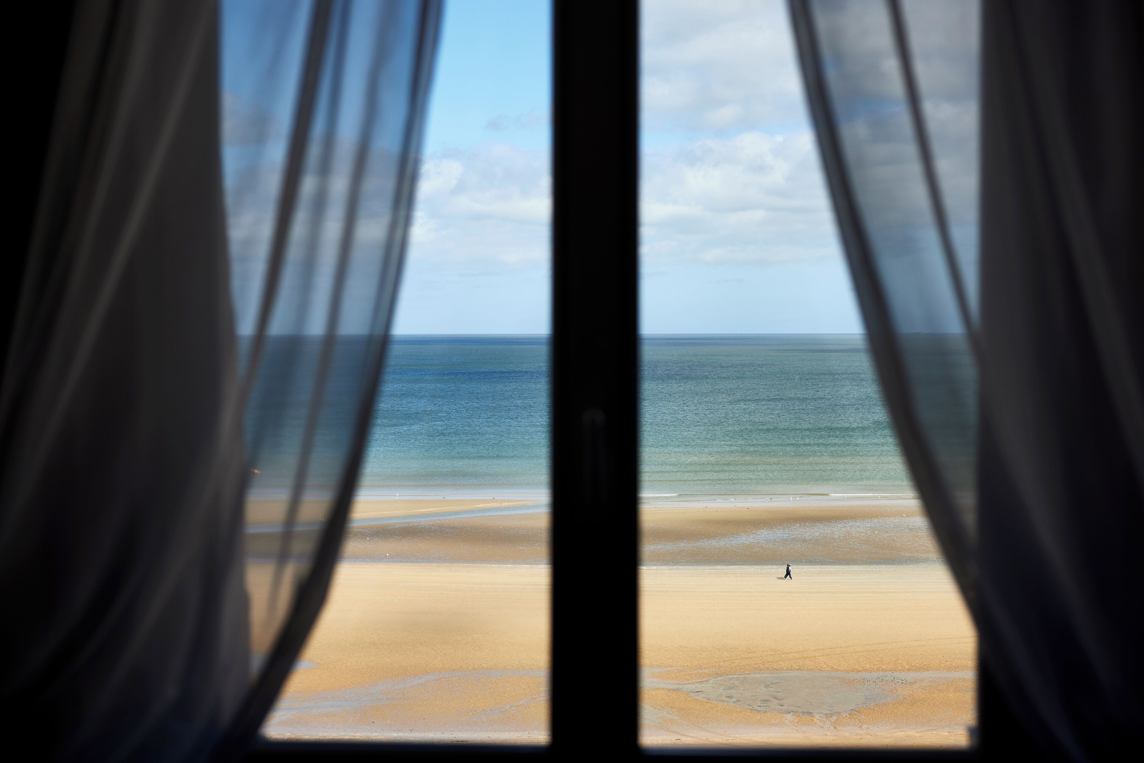 Vista de una playa a través de cortinas de tela translúcida, con una persona caminando sobre la arena.