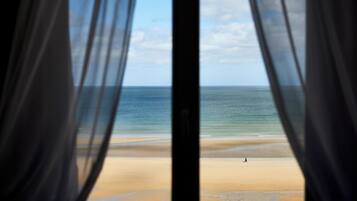Vista de una playa a través de cortinas de tela translúcida, con una persona caminando sobre la arena.