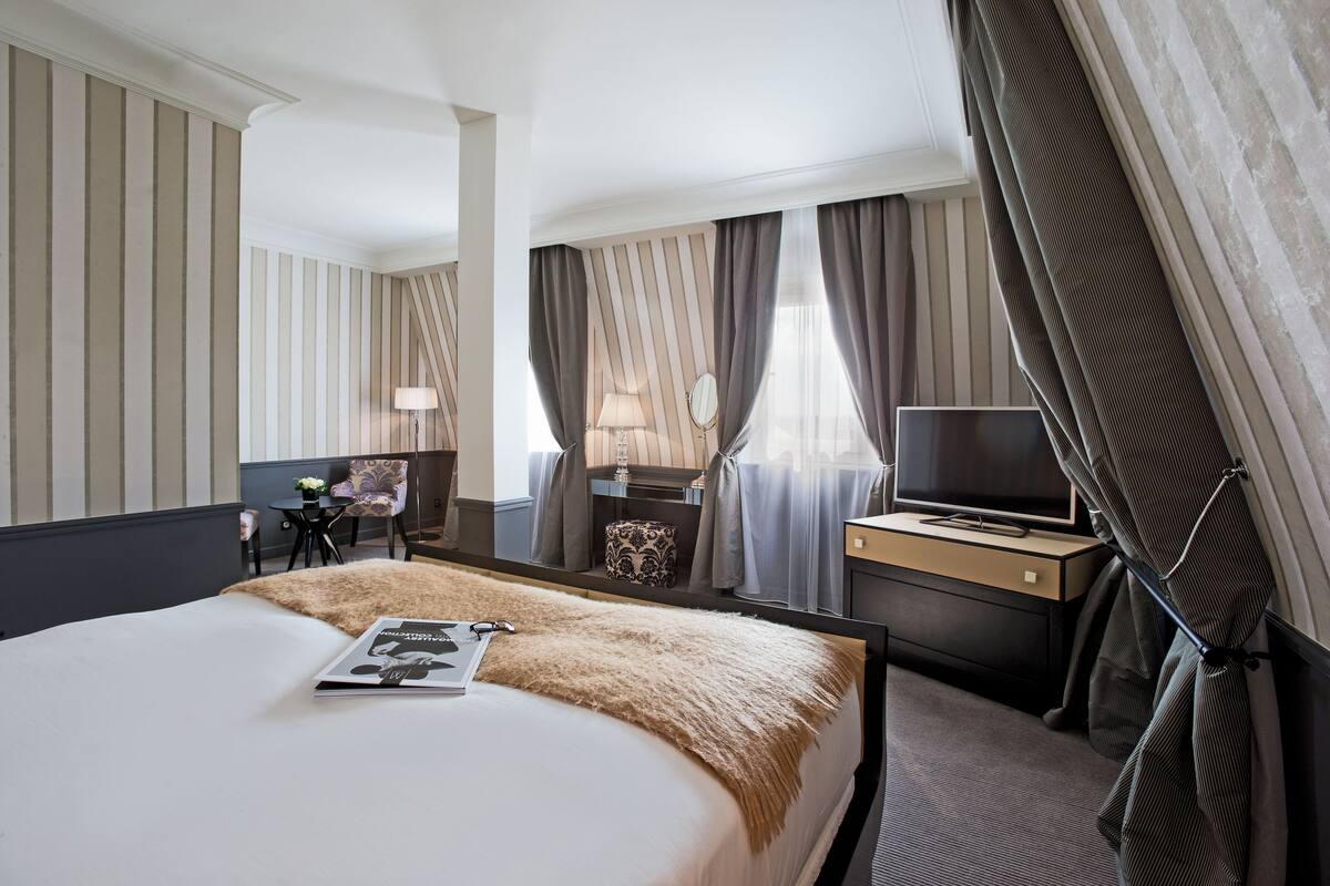 A hotel room with a large bed, a desk with a TV, a chair, and a small table with a lamp.