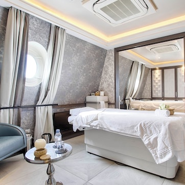A modern hotel room with a large bed, a sofa, a small round table with a lamp, and a window with curtains.