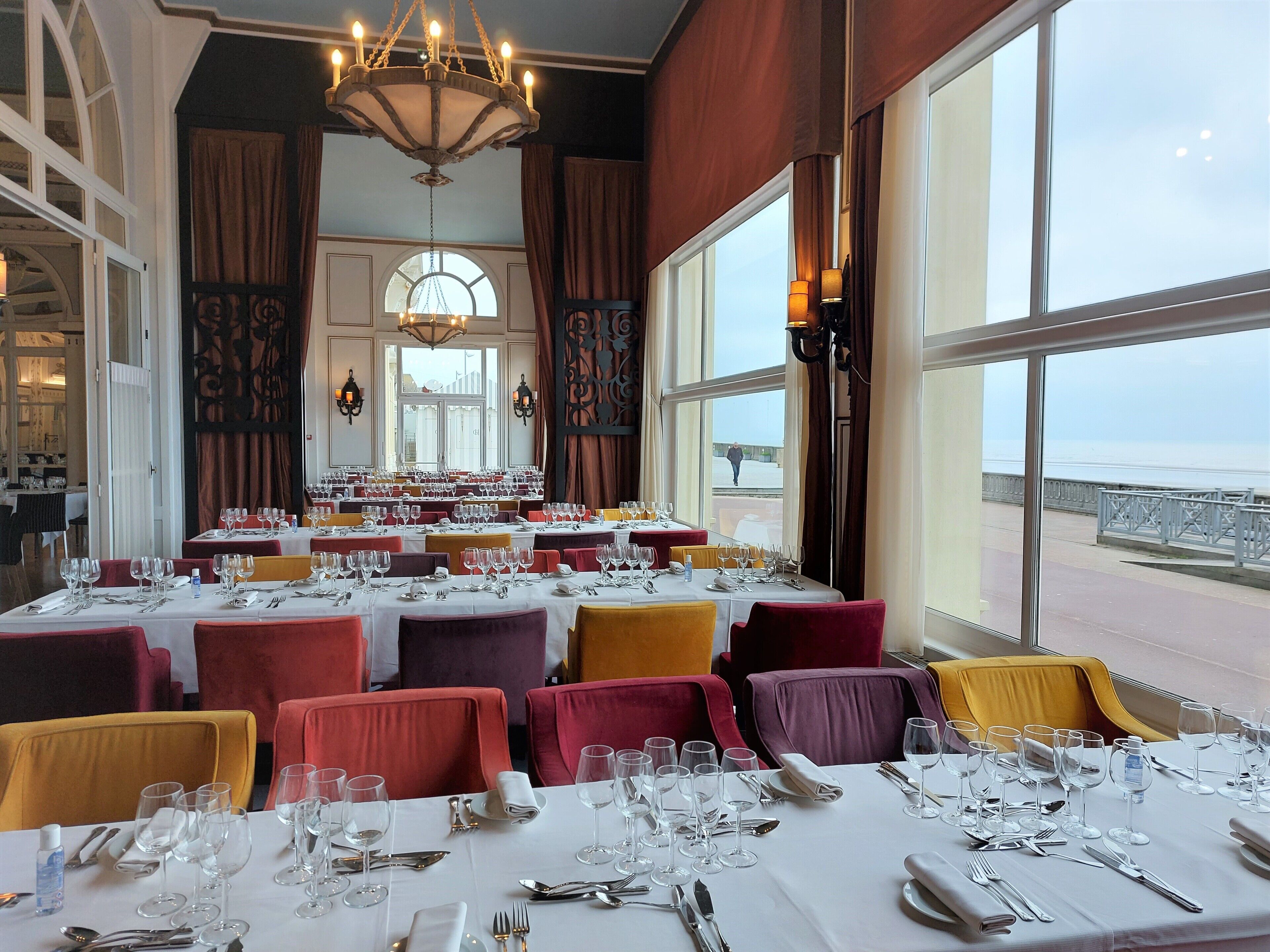 Un restaurant avec de nombreuses tables dressées, de grandes fenêtres donnant sur la mer et un lustre suspendu au plafond.