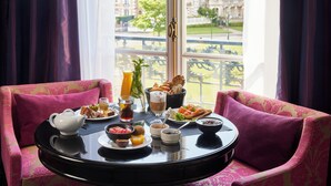 A breakfast table with a variety of foods and drinks, a window with a view of buildings, and two pink upholstered chairs. - Le Grand Hotel de Cabourg - MGallery Collection (Cabourg)