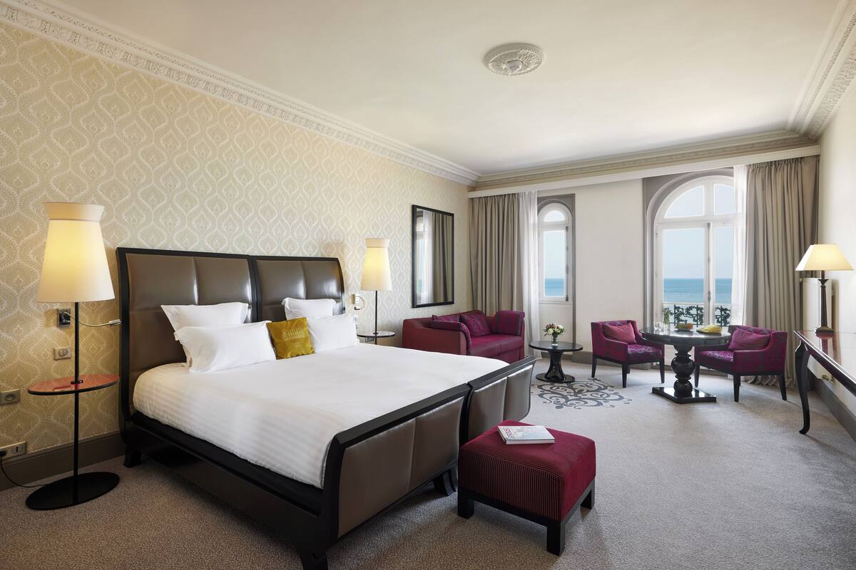 A hotel room with a large bed, two armchairs, a small table, and a view of the sea through large windows.