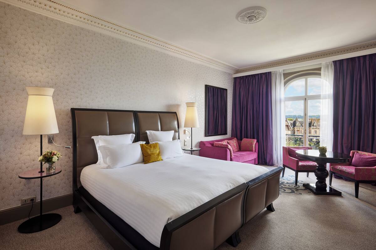 A hotel room with a large bed, two bedside lamps, a small table with a vase of flowers, a pink armchair, a round table, and a view of buildings through a window with purple curtains.