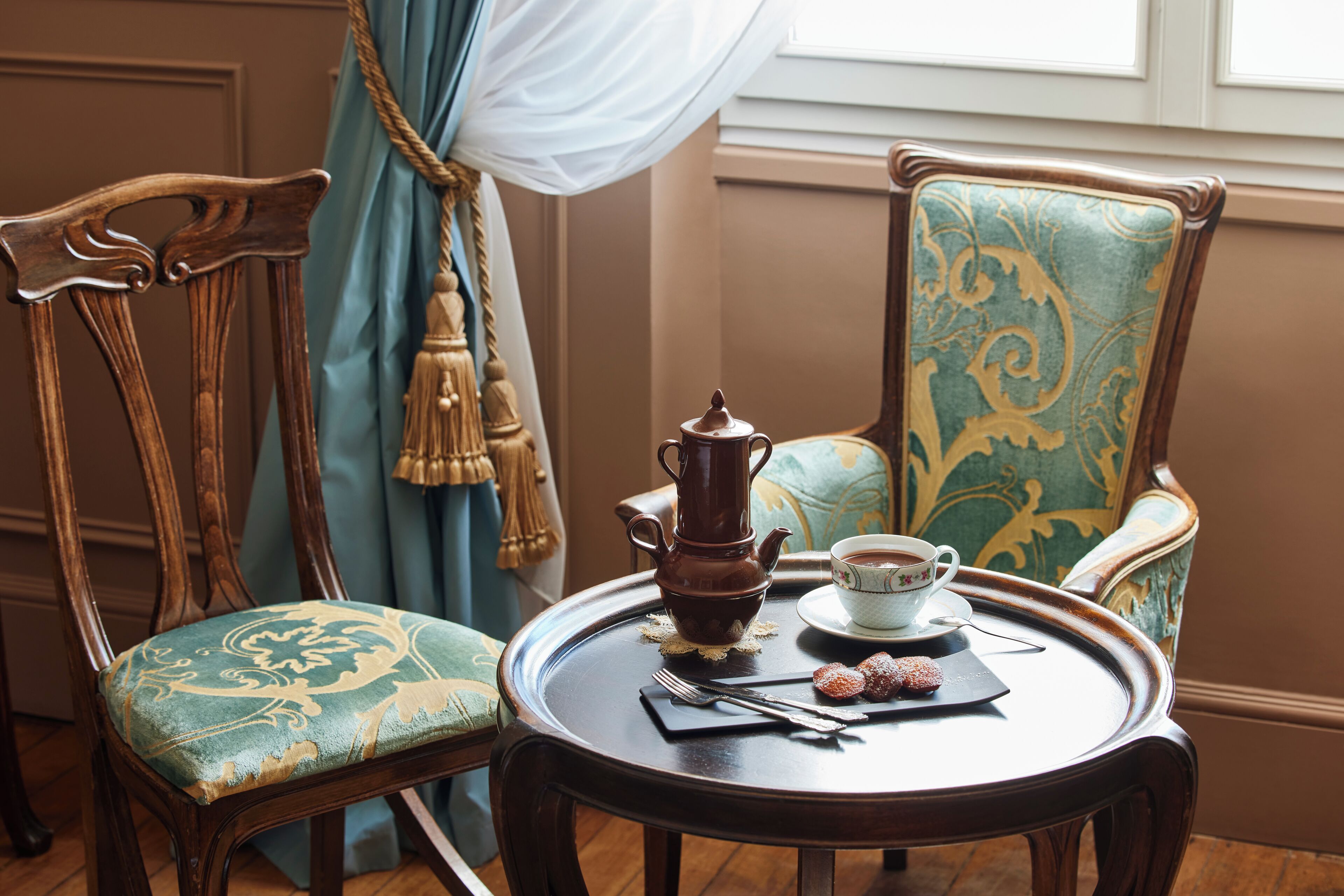 Une table à manger avec une théière, une tasse et des pâtisseries, placée entre deux chaises dotées d’un revêtement orné.