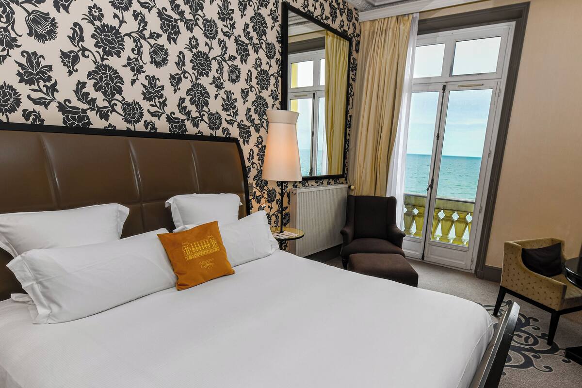 A hotel room with a bed, a chair, a table, and a view of the sea through large windows.
