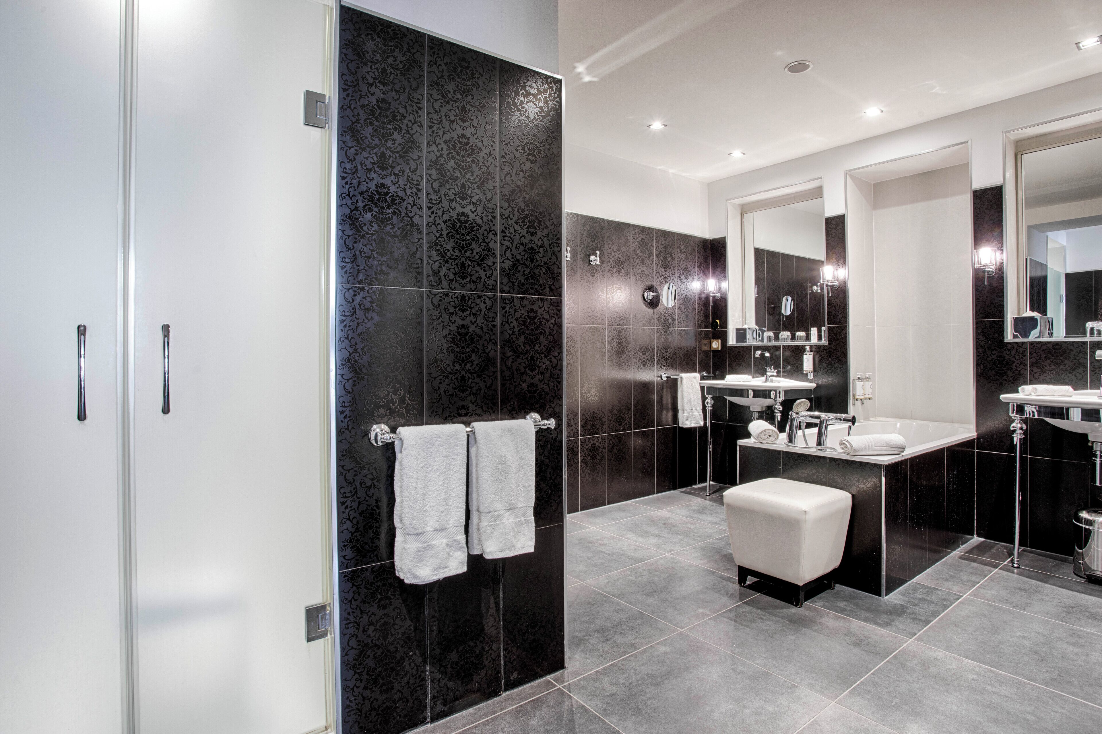 A modern bathroom with dark tiles, two sinks, a large mirror, and a white bench.