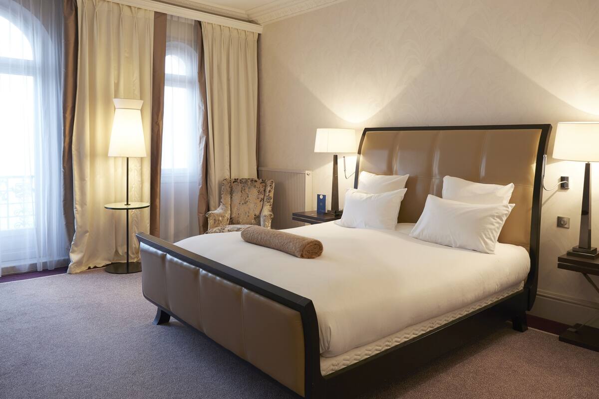A neatly made bed with a rolled towel, two bedside lamps, a chair, and a window with curtains.