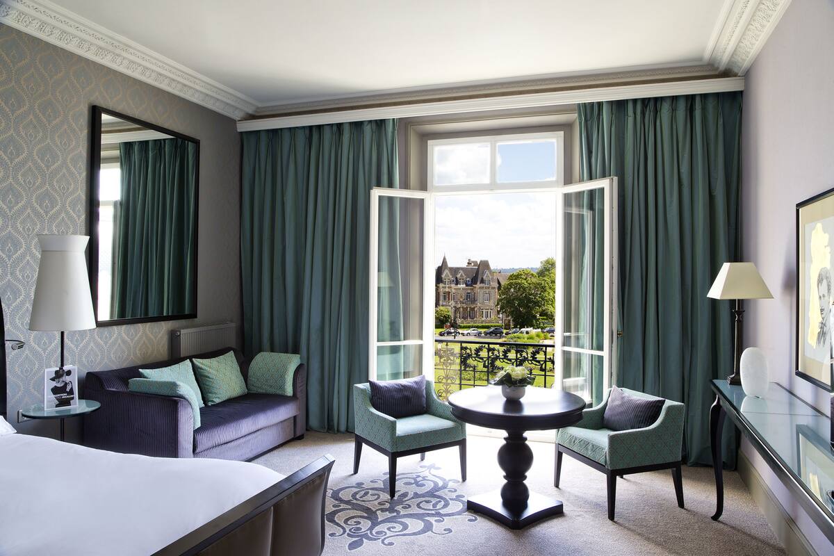 A hotel room with a large window, a sofa, two armchairs, a small table, and a bed.