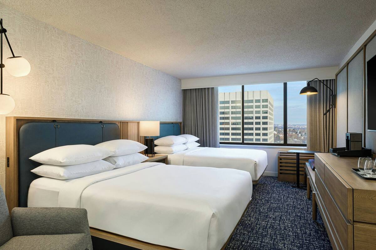 Habitación de hotel con dos camas, un escritorio, una silla, un televisor y un ventanal con vistas a la ciudad.
