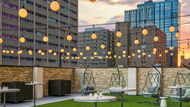 An outdoor seating area with artificial grass, tables, chairs, and hanging lights against a cityscape backdrop. - Sheraton Philadelphia University City Hotel (Philadelphia)