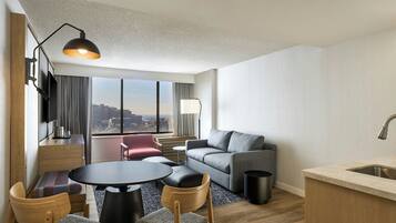 A modern hotel room with a grey sofa, a round dining table with chairs, a small kitchen area with a sink, and a city view through large windows.