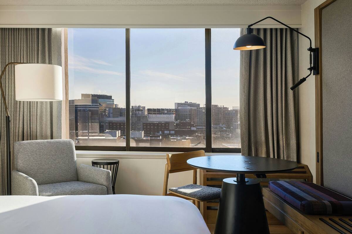 Habitación de hotel con cama, una silla, una mesa, un sofá, una lámpara y vistas de la ciudad a través de ventanas amplias.