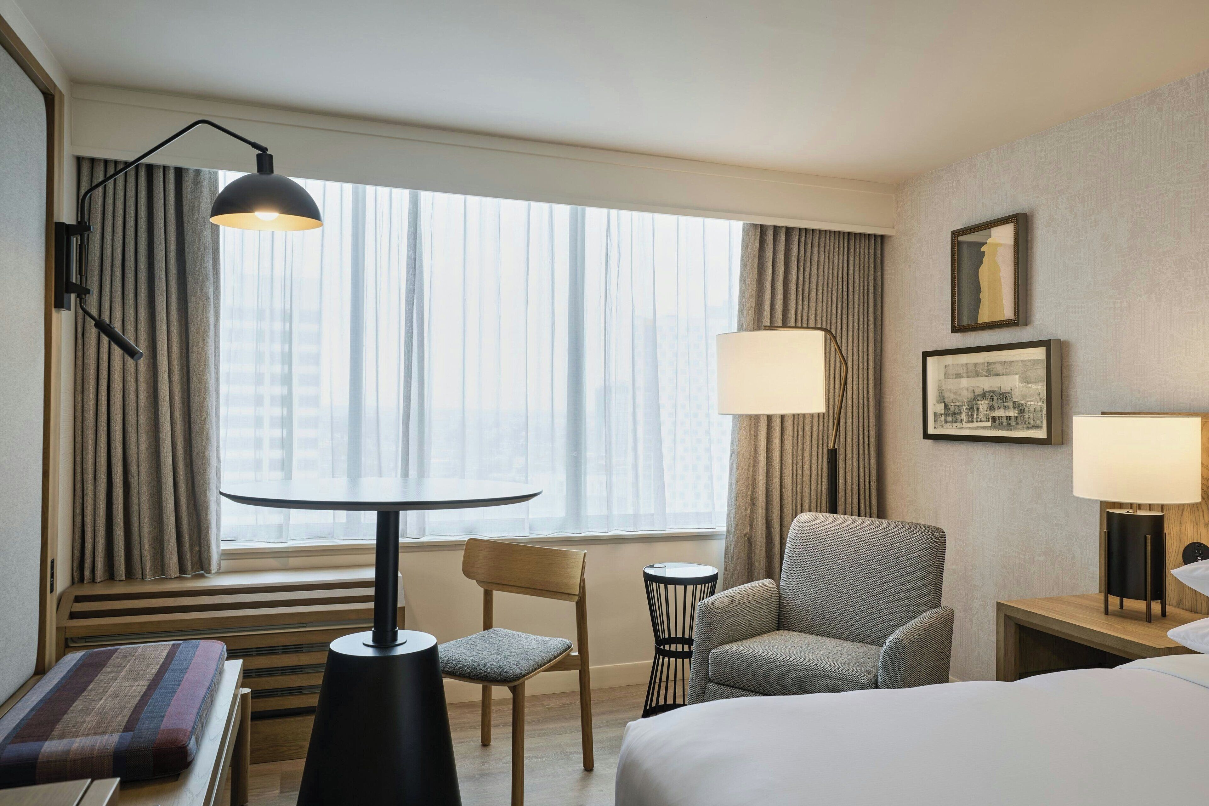 Ett hotellrum med en säng, ett skrivbord, en stol och ett fönster med gardiner.