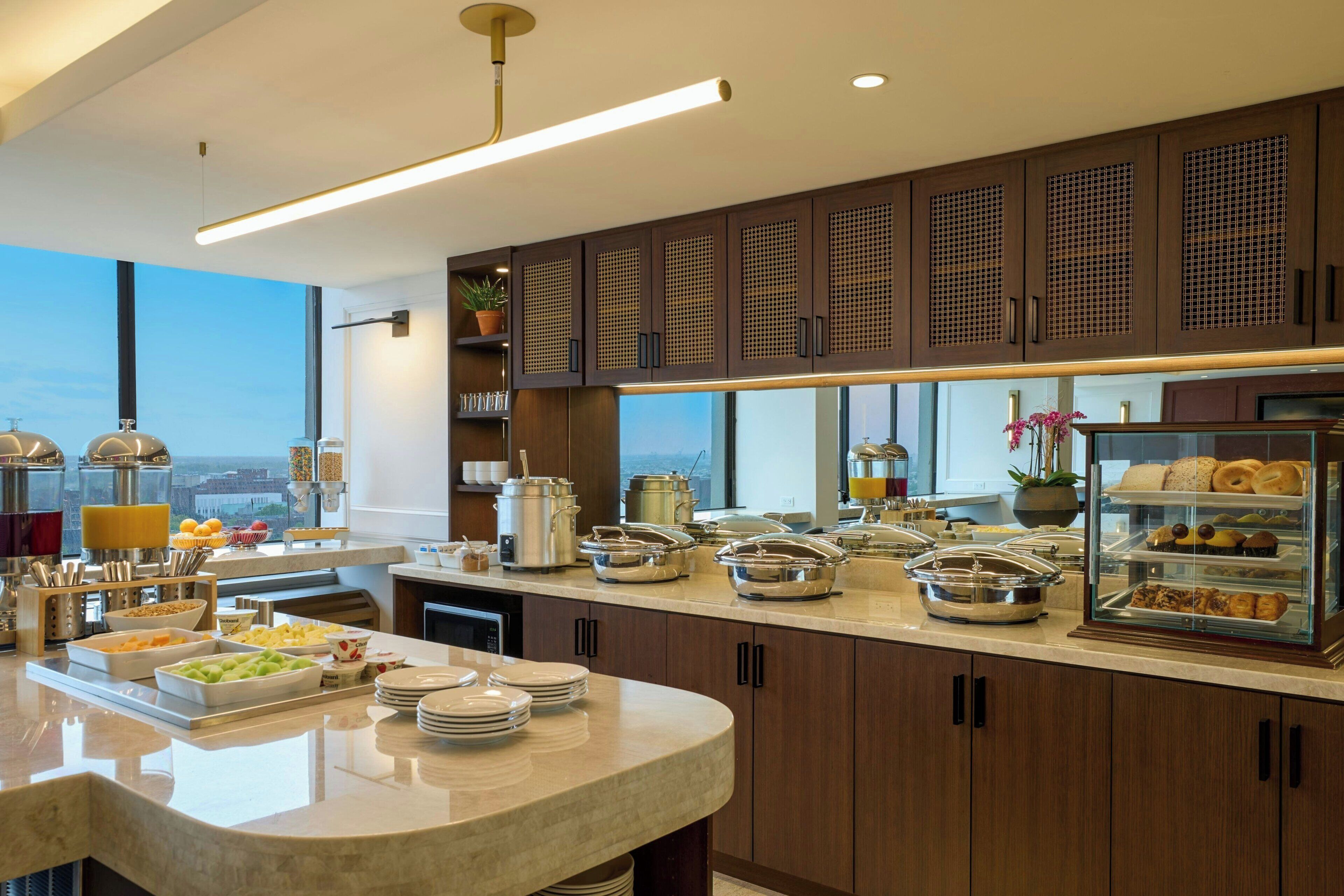 A modern kitchen with a marble countertop, buffet setup, and large windows offering a city view.